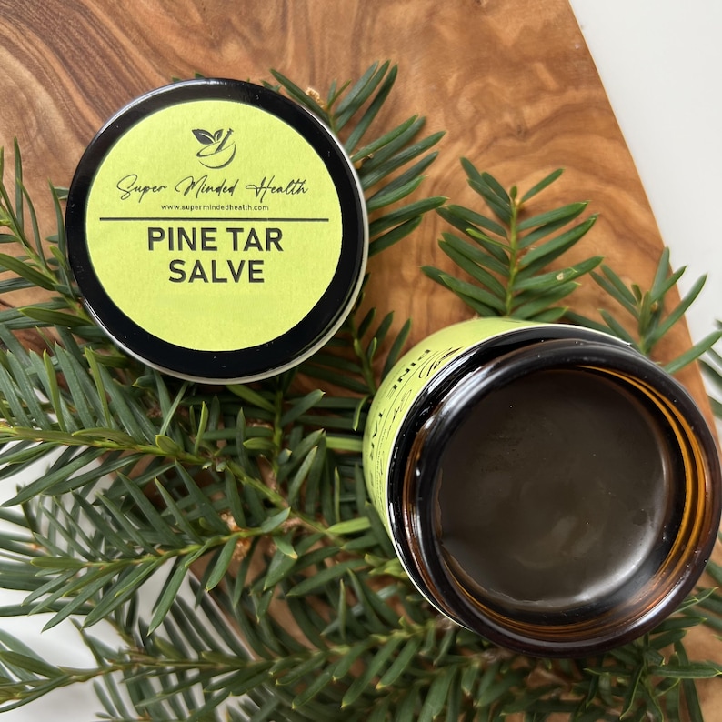 Puede incluir: Un recipiente redondo de b&aacute;lsamo de alquitr&aacute;n de pino con una etiqueta verde lima que muestra el texto Super Minded Health y Pine Tar Salve, junto a un tarro de vidrio &aacute;mbar abierto con ung&uuml;ento marr&oacute;n oscuro, sobre agujas de pino.