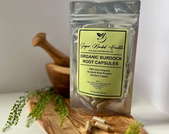 May include: A clear plastic bag filled with brown capsules labelled "Organic Burdock Root Capsules" with the brand name "Super Minded Health" and the website address "www.supermindedhealth.com". The bag is sitting on a wooden surface with green plants in the background.