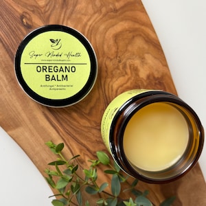 Op de afbeelding: Een klein potje met gele oregano-balsem met een zwart etiket waarop "Super Minded Health" en "OREGANO BALM" staat, met de tekst "Antifungal, Antibacterial, Antiparasitic" onder de productnaam. Het potje staat op een houten oppervlak met een klein groen plantje op de voorgrond.