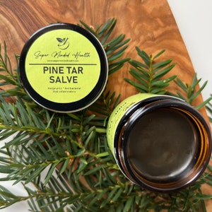 Può includere: Un barattolo verde con un coperchio nero e un'etichetta che recita "Super Minded Health" e "Pine Tar Salve". L'etichetta elenca anche i benefici del balsamo: "Antiprurito", "Antibatterico" e "Antinfiammatorio". Il barattolo è appoggiato su una superficie di legno con vegetazione verde.
