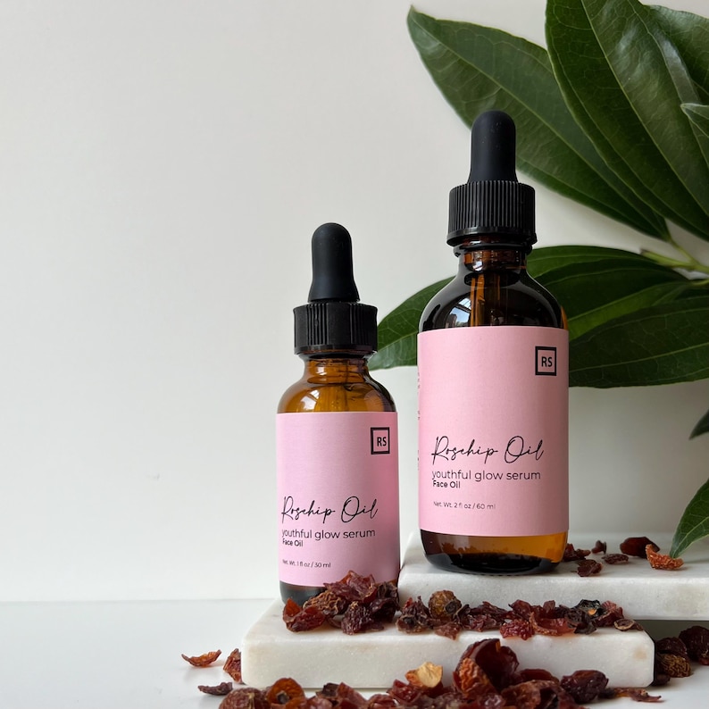 May include: Two amber glass bottles with dropper tops, labelled "Rosehip Oil youthful glow serum Face Oil" with the text "RS" in a square on the label. The bottles are on a white surface with dried rosehips.