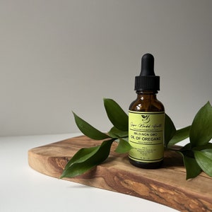 May include: A brown glass bottle with a black dropper top, labeled "Super Minded Health" and "Wild-Non GMO Oil of Oregano" on a wooden surface with green leaves.