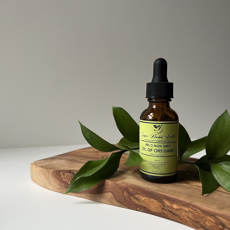 May include: A brown glass bottle with a black dropper top, labeled "Super Minded Health" and "Wild-Non GMO Oil of Oregano" on a wooden surface with green leaves.