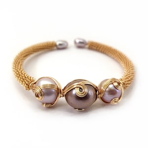 May include: Gold-toned wire bracelet featuring three large, iridescent pearls. The pearls are wrapped in gold wire, and the bracelet has a woven gold band. The pearls are a mix of lavender and taupe colors.