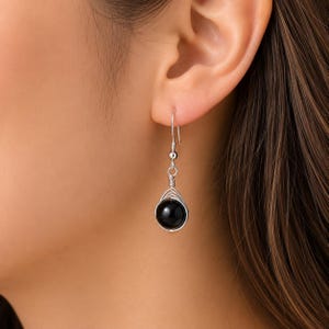 May include: A silver-toned earring featuring a smooth, round black bead encased in a delicate wire-wrapped cage design. The earring hangs from a simple silver-toned hook, showcasing a polished finish and a minimalist structure that dangles.