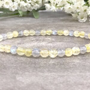May include: A delicate bracelet made with small, round beads of yellow and light blue gemstones. The bracelet is laid out on a gray textured surface.