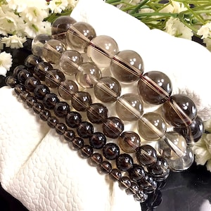 May include: Five stacked bracelets made of smoky quartz beads. The bracelets are made of different sizes of round beads, with the largest beads at the top and the smallest beads at the bottom. The beads are a dark brown color with a slight shimmer.