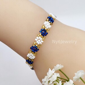 May include: A beaded bracelet with a floral pattern. The bracelet features white, gold, and dark blue beads. The bracelet is shown on a wrist. The text "Ny6Jewelry" is visible on the wrist.