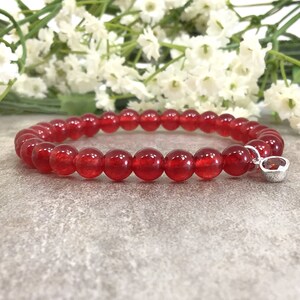 May include: Red gemstone bracelet with a silver charm featuring a smaller red gemstone. The bracelet is made of round beads.