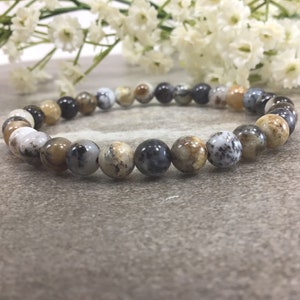 May include: A beaded bracelet featuring a variety of earth-toned, round stones. The beads are primarily shades of brown, gray, and cream, with unique patterns and textures. The bracelet is displayed on a neutral surface, with white flowers in the background.