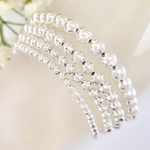 May include: A set of five silver bead bracelets, each with a different size bead. The bracelets are stacked on top of each other, creating a layered look.