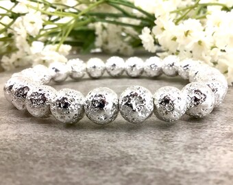 May include: A silver beaded bracelet with textured, spherical beads. The bracelet is strung together, and the beads have a rough, metallic appearance. The jewelry is displayed against a neutral background with white flowers.