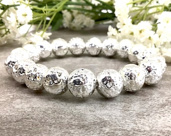 May include: A silver beaded bracelet with textured, spherical beads. The bracelet is made of multiple round beads with a hammered or pitted surface. The beads are strung together to form a complete circle. The background includes white flowers and greenery.