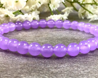 May include: A bracelet made of smooth, round, lavender-colored beads. The beads are strung together to form a flexible band. The bracelet is displayed on a gray surface, with white flowers in the background.
