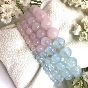 May include: Two beaded bracelets, one pink and one light blue, are stacked on a white surface. The bracelets are made of round beads.