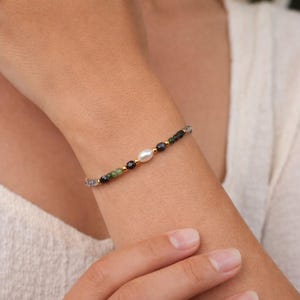 May include: A delicate bracelet featuring a central white pearl, accented by small black, green, and gold-colored beads. The bracelet is strung on a fine gold chain, creating an elegant and understated piece of jewelry.