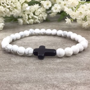May include: White beaded bracelet with a black cross charm. The bracelet is made of small, round beads and has a simple design.