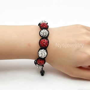 May include: A bracelet featuring alternating red and clear crystal-encrusted spherical beads strung on a black cord. The design includes a drawstring closure with two small black beads at the ends. The text Ny6Jewelry is visible on the right side.