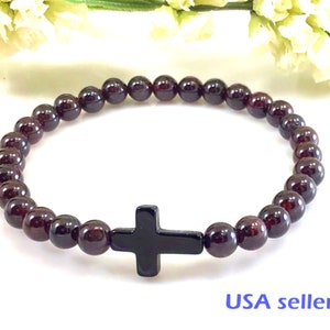 May include: A red garnet gemstone bracelet with a black cross charm. The bracelet is made with round beads and has a simple design.