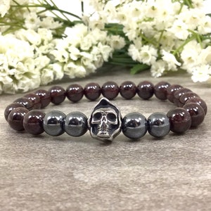 May include: A beaded bracelet featuring dark red and gray beads with a silver skull charm. The bracelet is made of polished, round beads. The skull charm is detailed with a hood.