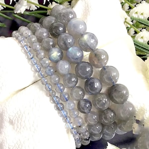 May include: A set of five grey labradorite gemstone bracelets with iridescent blue flashes. The bracelets are stacked on top of each other and are made with round beads.