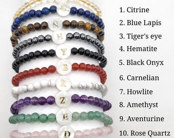 May include: A stack of ten beaded bracelets in various colors, including yellow, blue, brown, black, red, white, purple, green, and pink. Each bracelet has a round, white charm with a gold letter.