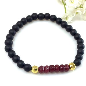 May include: A black beaded bracelet with a row of red gemstone beads and gold accents.