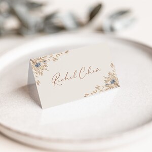 May include: A white place card with a gold and blue floral design. The name "Rachel Cohen" is written in cursive on the card.