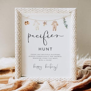 May include: A white framed sign with the words "pacifier HUNT" in elegant script. The sign includes text about a game and is decorated with baby clothes and pacifiers. The sign is on a wooden tray with a beige cloth.
