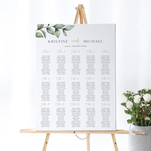 May include: A white wedding seating chart with green foliage and gold lettering. The chart lists the names of guests assigned to each table number. The chart is displayed on a wooden easel.