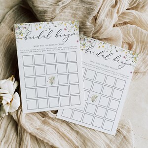 May include: Two floral bridal bingo cards with a white background and black grid lines. The text "Bridal Bingo" is written in cursive at the top of each card. The text "What will the bride receive?" is written below the title. The cards are on a beige fabric background.