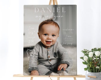 May include: A white and grey first birthday sign featuring a photo of a baby boy. The sign includes the baby's name, Daniel, and details about his first year, such as his weight, height, and favourite things.