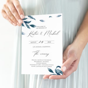 Puede incluir: Un programa de boda blanco con detalles florales en acuarela azul es sostenido. El programa incluye los nombres "Kristine & Michael", la fecha 24 de agosto de 2022 y la ubicación Los Ángeles, California.