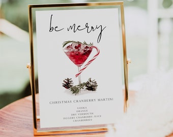 May include: A gold framed sign with the text "be merry" and a watercolor illustration of a Christmas cranberry martini. The recipe is listed below the illustration: "Christmas Cranberry Martini, Vodka, Orange, Dry Vermouth, Jiggers Cranberry Juice, Cranberries."