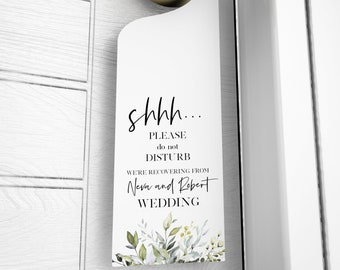 Puede incluir: Colgador de puerta blanco con pomo dorado. El letrero dice "shhh... PLEASE do not DISTURB WE'RE RECOVERING FROM Neva and Robert WEDDING" con detalles florales. El letrero está colgado en una puerta.