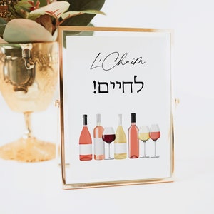 May include: A gold-framed sign with the text "L'Chaim! לחיים!" in black and white. The sign features a row of six wine bottles and two wine glasses, all in various shades of pink, red, and white.