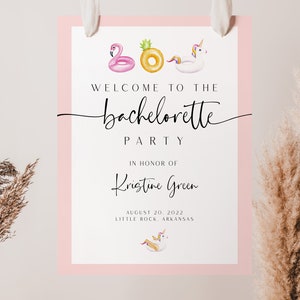May include: A white sign with a pink border announcing a hen party for Kristine Green on 20 August 2022 in Little Rock, Arkansas. The sign features illustrations of inflatable pool floats, including a flamingo, pineapple, and unicorn.