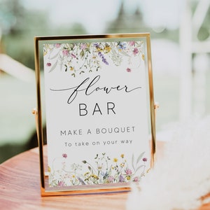 Pu&ograve; includere: Un cartello con cornice dorata con le parole "Flower Bar" in elegante scrittura. Il cartello recita anche "Make a Bouquet To take on your way". Il cartello &egrave; decorato con illustrazioni floreali ad acquerello e poggia su una superficie di legno.