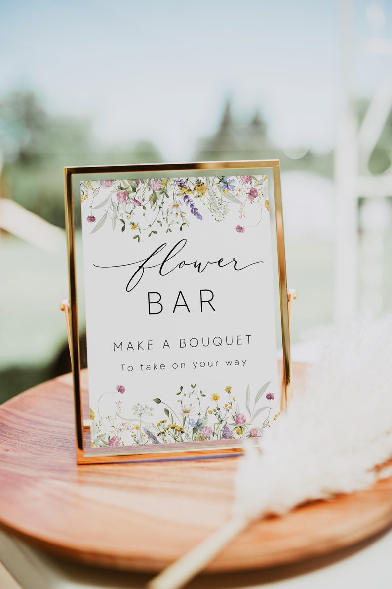 Pu&ograve; includere: Un cartello con cornice dorata con le parole "Flower Bar" in elegante scrittura. Il cartello recita anche "Make a Bouquet To take on your way". Il cartello &egrave; decorato con illustrazioni floreali ad acquerello e poggia su una superficie di legno.