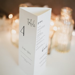 Op de afbeelding: Witte tafelnummerkaart met zwarte kalligrafie voor tafelnummer 4. De kaart bevat ook de namen Kristine + Michael en de datum 8.29.2023. De kaart bevat ook een menu met de koppen "Voorgerecht", "Hoofdgerecht", "Nagerecht" en "Drankjes".