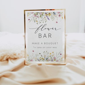 Pu&ograve; includere: Un cartello con cornice dorata con le parole "Flower Bar" in elegante scrittura, che invita gli ospiti a "Make a Bouquet". Il cartello &egrave; adornato da un bordo floreale di fiori selvatici e poggia su una superficie satinata.