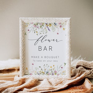 Pu&ograve; includere: Un cartello incorniciato con le parole "Flower Bar" e "Make a Bouquet" in elegante scrittura. Il cartello &egrave; decorato con illustrazioni floreali ad acquerello e posto su un vassoio di legno con un tessuto strutturato.