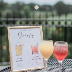 May include: A framed sign with the title "Signature Drinks" lists two cocktails: "Dr. J's Painkiller" and "Coach K's Gym and Tonic". The sign is next to two glasses of cocktails, one yellow and one pink.