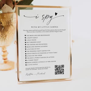 May include: A framed wedding game card with the words "i spy" at the top. The card includes a list of photo prompts and instructions. The frame is gold-colored, and the text is in black.