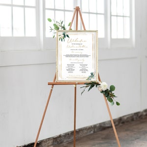 May include: A gold-framed wedding welcome sign with greenery and white flowers. The sign includes the text "Welcome to the wedding of Kristina & Matthew" and the date "August 21, 2023 | 1:00 PM".