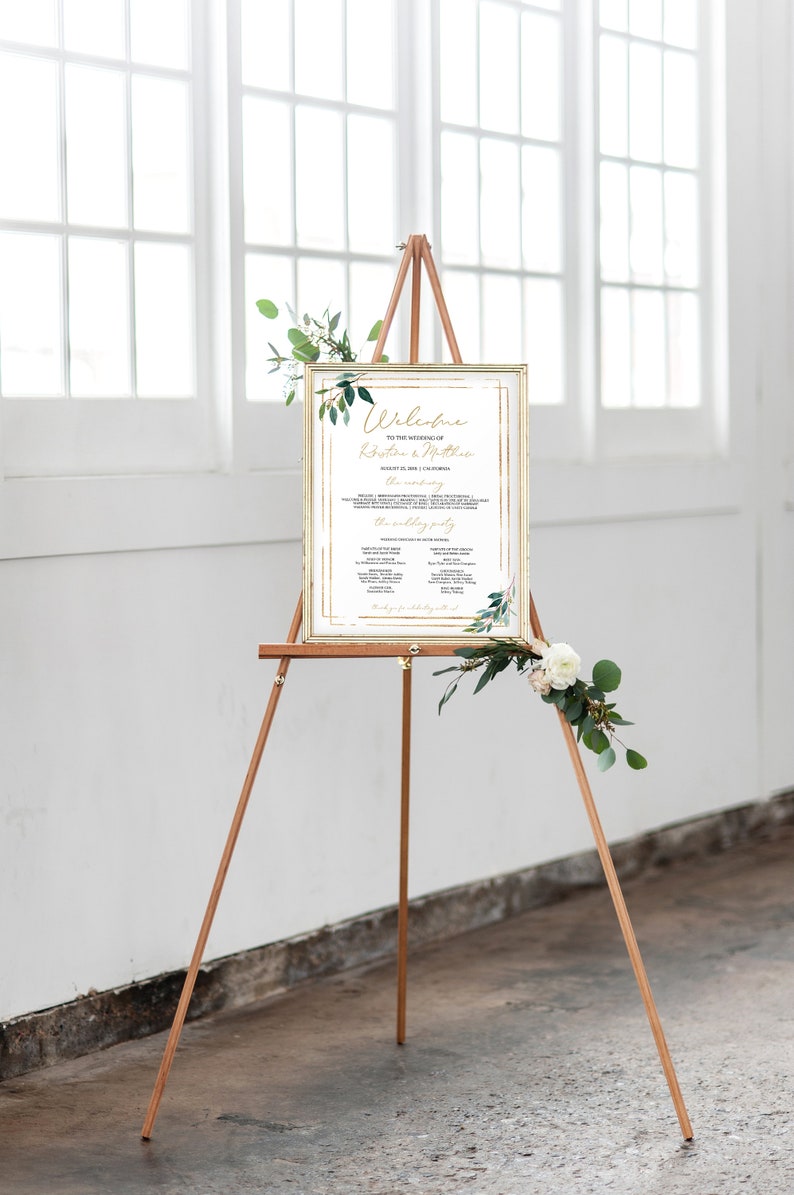 May include: A gold-framed wedding welcome sign with greenery and white flowers. The sign includes the text "Welcome to the wedding of Kristina & Matthew" and the date "August 21, 2023 | 1:00 PM".