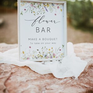 Pu&ograve; includere: Un cartello incorniciato bianco con la scritta "Flower Bar" in corsivo, che invita gli ospiti a "Make a Bouquet". Il cartello &egrave; decorato con illustrazioni floreali e poggia su una superficie di pietra con un tessuto bianco drappeggiato.