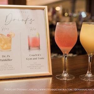 May include: A gold framed sign with the text "SIGNATURE Drinks" and two cocktail recipes: "Dr. J's Painkiller" with ingredients rum, orange juice, pineapple juice, and cream of coconut, and "Coach K's Gym and Tonic" with ingredients gin, tonic water, pomegranate juice, and a hint of lime. Two glasses of cocktails are on the bar counter, one pink and one yellow.