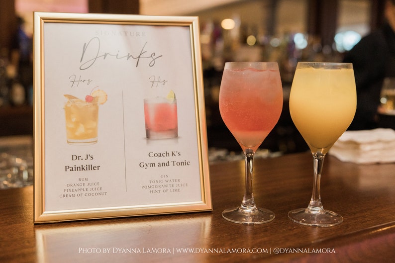 May include: A gold framed sign with the text "SIGNATURE Drinks" and two cocktail recipes: "Dr. J's Painkiller" with ingredients rum, orange juice, pineapple juice, and cream of coconut, and "Coach K's Gym and Tonic" with ingredients gin, tonic water, pomegranate juice, and a hint of lime. Two glasses of cocktails are on the bar counter, one pink and one yellow.