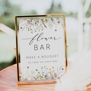 Pu&ograve; includere: Un cartello con cornice dorata con le parole "Flower Bar" in elegante scrittura, che invita gli ospiti a "Make a Bouquet". Il cartello &egrave; decorato con un bordo floreale di fiori selvatici nei toni del rosa, giallo e viola. Il cartello &egrave; su un tavolo di legno.